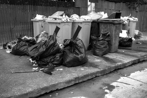 A collection of black plastic rubbish bags filled with waste sits on a concrete pavement next to a row of large wheelie bins in an outdoor area. The bins are filled with various discarded items, including cardboard boxes, paper, and plastic packaging, and are positioned against a wooden fence. Some of the bags and waste are leaning against the bins, while others are stacked on the ground, with a few pieces of rubbish scattered nearby. The scene appears to be part of a rubbish removal or clearance process, typical of urban residential waste collection in Leytonstone, with the environment being a typical local street or yard setting. Leytonstone House Clearance often handles such rubbish removal tasks in the postcode area, providing professional services for clearing waste from households or properties. The lighting is natural and even, highlighting the different textures of the materials and the arrangement of the waste items.