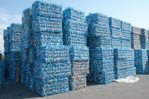 A large outdoor storage area featuring numerous tightly packed stacks of clear plastic bottles, primarily blue and transparent, arranged in rectangular blocks on a paved surface. Some bottles have slight variations in shades of blue and clear, with a few scattered bottles and plastic debris on the ground nearby. The background shows a clear, bright sky with no visible structures or vegetation, indicating an industrial or waste management yard likely associated with rubbish collection or recycling services in the Leytonstone area. The scene is well-lit, with natural daylight highlighting the textures and shapes of the compressed bottles, representing the type of waste material processed during rubbish removal operations carried out by Leytonstone House Clearance or similar local services.