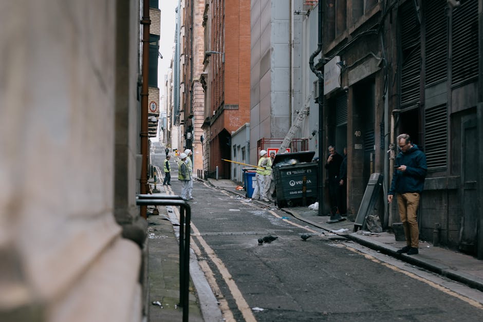 A narrow urban street scene showing a group of construction or maintenance workers in high-visibility clothing and safety helmets, gathered near a black waste bin with an open lid, surrounded by scatt